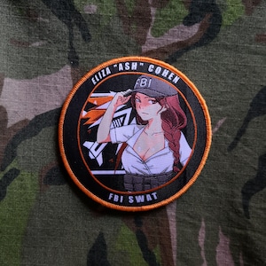 May include: A black and orange embroidered patch with a cartoon woman wearing a white shirt, black cap, and a black belt with the text "FBI SWAT" on it. The text "ELIZA "ASH" COHEN" is around the outside of the patch.