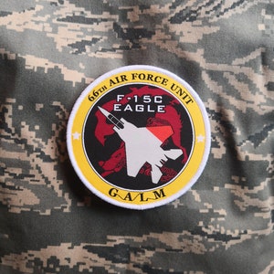 May include: A yellow and white patch with the text "66th Air Force Unit" and "F-15C Eagle" in black. The patch also features a white silhouette of an F-15C Eagle fighter jet against a red and black background. The text "G/A/L/M" is printed in black at the bottom of the patch.