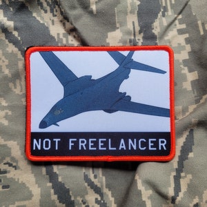 May include: A black and white patch with a silhouette of a bomber plane and the text "NOT FREELANCER" in white letters on a black background. The patch has a red border.
