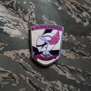 May include: A beige patch with a purple and white helmet with a pink wing. The patch has the text "51st Tactical Fighter Sq" and "INVIDIOUS" in a red banner.