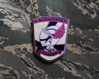 May include: A beige patch with a purple and white helmet with a pink wing. The patch has the text "51st Tactical Fighter Sq" and "INVIDIOUS" in a red banner.