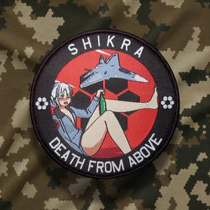 May include: A circular embroidered patch featuring a red background with a black honeycomb pattern. It depicts an anime-style character holding a green bottle, with a fighter jet above. The text reads SHIKRA and DEATH FROM ABOVE in white.