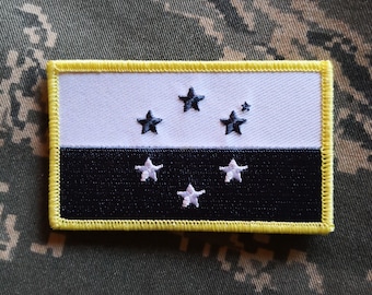 May include: A black and white embroidered patch with five white stars and a yellow border. The patch is on a camouflage fabric background.