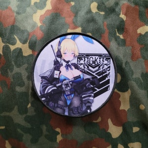 May include: A round, white patch with a black border features an anime-style illustration of a woman with blonde hair, blue eyes, and a blue bunny ear headband. She is holding a rifle and wearing a black jacket, blue shirt, and black pants. The patch also includes a black and white logo with the letters "G.K."