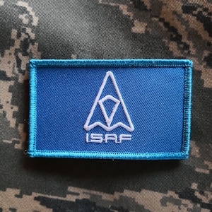 Op de afbeelding: Een blauwe patch met een witte gestileerde driehoek en de tekst "ISAF" in wit.