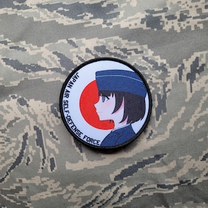 Könnte beinhalten: Ein runder Aufnäher mit einer Anime-Illustration einer Person in blauer Uniform und Hut, vor einem roten Kreis und weißem Hintergrund. Der Text "JAPAN AIR SELF-DEFENSE FORCE" ist am Rand aufgedruckt. Der Aufnäher befindet sich auf einem Tarnstoff.