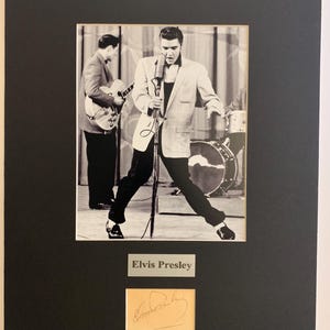 May include: Black and white framed photograph of Elvis Presley performing on stage, holding a microphone. The image includes a label that reads "Elvis Presley" and a signature.