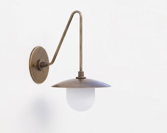 May include: A wall-mounted light fixture featuring a circular brass backplate, an angled brass arm, a wide brass shade, and a spherical frosted white glass globe. The metal components have a matte finish and the design is simple and functional.