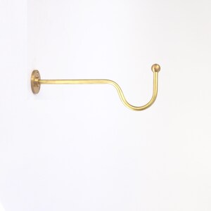 May include: A brass-colored wall-mounted coat hook. The hook has a circular mounting plate with screw holes, a straight horizontal bar, and a curved hook shape ending in a small ball. The hook is mounted on a white wall.