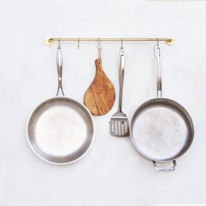 May include: A brass kitchen utensil rack with three hanging silver pans and a wooden cutting board.