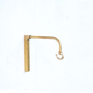 May include: A brass wall-mounted hook with a curved arm and a hook at the end. The hook is attached to a square base.