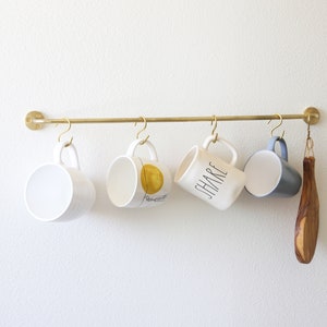 May include: A brass-colored metal bar with four white ceramic mugs hanging from it. One mug has a gold design, another says "SHARE", and the last is blue inside. A wooden spoon hangs on the right.