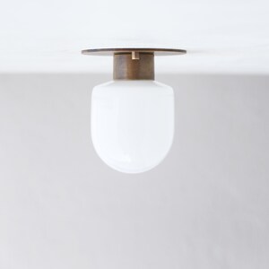 May include: A modern ceiling light fixture with a white, rounded glass shade. The fixture has a brass-colored base and a circular wooden top. The light is designed to be mounted on a ceiling.
