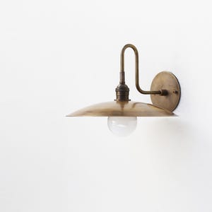 May include: A wall-mounted light fixture featuring a circular brass backplate, a curved brass arm, and a wide, flat, saucer-shaped brass shade. A single, spherical, frosted white glass light bulb is positioned beneath the center of the shade.