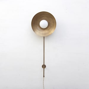 May include: A brass wall sconce with a circular shade and a white globe bulb. The sconce has a long, thin arm and a small, round base. The fixture is mounted on a white wall.