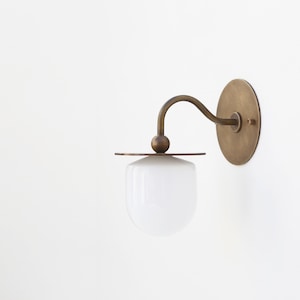 May include: A brass wall sconce with a white glass shade. The sconce has a curved arm and a round base.