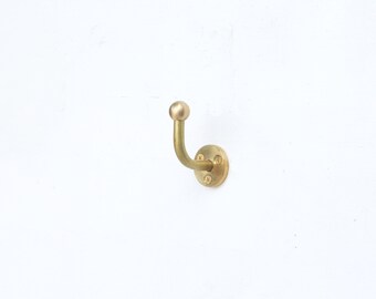 May include: A brass wall hook with a rounded ball top and a curved hook design. The hook is attached to a circular base with four screws. The brass finish gives the hook a vintage or antique look.