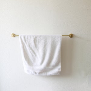 Puede incluir: Una toalla blanca cuelga de una barra de toalla de metal dorado montada en una pared blanca.