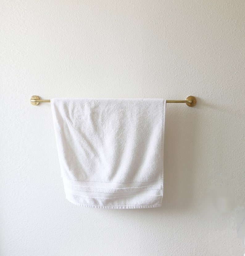 Puede incluir: Una toalla blanca cuelga de una barra de toalla de metal dorado montada en una pared blanca.