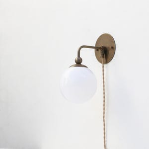 May include: A wall-mounted light fixture with a round, white glass globe. The fixture has a brass-colored metal arm and a circular backplate. A braided, tan-colored cord hangs down from the fixture.
