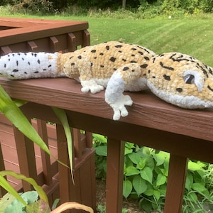 May include: A plush, handmade leopard gecko toy. The reptile is beige with black spots and a white belly and tail. It is lying on a brown wooden deck railing with green foliage in the background. The gecko has white feet and a white head.