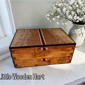 May include: A handcrafted wooden box with two drawers, featuring a natural wood grain finish. The box has a dark border and a decorative stripe down the center. The text "Little Wooden Hart" is at the bottom.