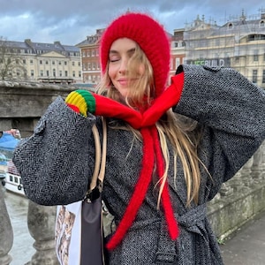 May include: A bright red fuzzy knit beanie and a matching red scarf with long fringe ends. The scarf features green and yellow striped accents at the ends. The set is worn with a dark gray herringbone patterned coat and a white tote bag.