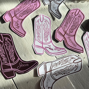 May include: Pink and white cowboy boot shaped place cards with names written on them. The names are: Jess, Bab, Hannah, Joan, Joan, and Allison.