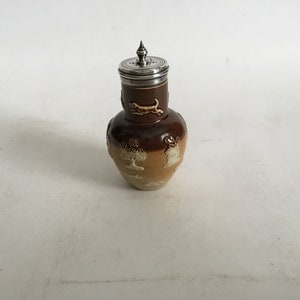 May include: A brown and cream ceramic jug with a silver lid. The jug has a hunting scene design with trees and a dog.
