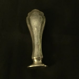 Op de afbeelding: Een zilveren zegel met een decoratief, versierd ontwerp. De zegel heeft een ronde basis en een hoog, slank lichaam.