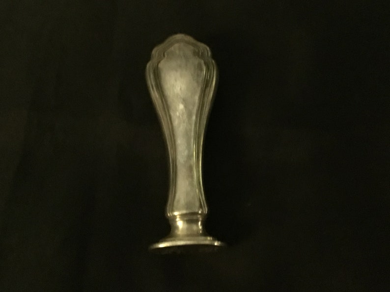 Op de afbeelding: Een zilveren zegel met een decoratief, versierd ontwerp. De zegel heeft een ronde basis en een hoog, slank lichaam.