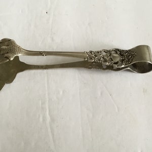 Puede incluir: Pinzas de servir de plata con un diseño decorativo y una pata de pájaro en un extremo. Las pinzas están marcadas con "Sterling" en la parte posterior.