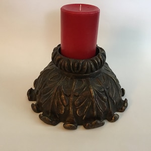 Puede incluir: Un portavelas con un diseño decorativo y ornamentado. La base es de metal y tiene un acabado marrón oscuro. El portavelas presenta un borde circular elevado y un patrón de hojas detallado. Una vela roja se coloca en la parte superior del portavelas.