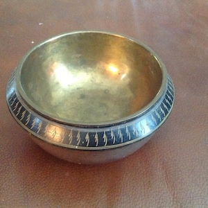 Könnte beinhalten: Eine kleine, runde Schale aus Messing mit einem schwarzen und goldenen Rand. Die Schale hat ein dekoratives Muster aus kleinen, stilisierten Bäumen um den Rand.