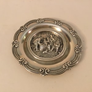 May include: A silver-plated dish featuring a raised design of three figures in a scene. The dish has a scalloped edge with a decorative pattern.