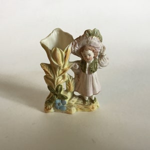May include: A porcelain figurine of a girl wearing a pink dress and a hat, holding a flower. The figurine is attached to a vase shaped like a flower.