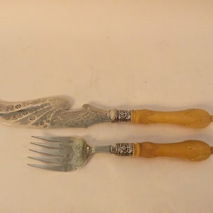 Puede incluir: Un juego de cubiertos de plata con un cuchillo y un tenedor. Los mangos están hechos de marfil y tienen un diseño detallado. La hoja del cuchillo es adornada y tiene un patrón decorativo.