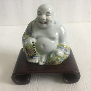 Puede incluir: Una figura de Buda sonriente, sentado, con cuerpo blanco y ropa estampada. La estatua de cerámica está sobre un soporte de madera marrón oscuro. La ropa del Buda presenta diseños amarillos, azules y florales.