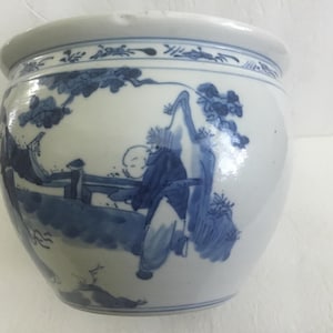 Könnte beinhalten: Ein weißer Keramik-Pflanztopf mit einem blau-weißen, asiatisch inspirierten Design. Der Pflanztopf hat eine abgerundete Form mit einem breiten Rand und ist mit einer detaillierten Szene von Figuren und Laub verziert.