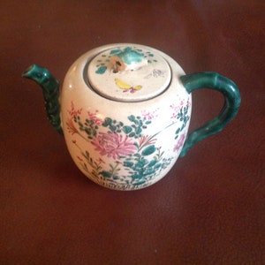 Könnte beinhalten: Eine weiße Teekanne aus Keramik mit einem grünen Henkel und Deckel. Die Teekanne ist mit floralen Mustern in Rosa, Grün und Gelb verziert.