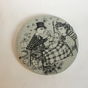 May include: A black and white illustration of a family with a baby. The family is dressed in festive attire and the baby is being presented with a gift. The illustration is on a round, white ceramic plate with a green background.