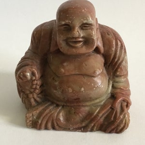 Puede incluir: Pequeña estatua de Buda marrón rojizo con una expresión sonriente. La figura está sentada, con una barriga redonda y túnicas detalladas. La estatua parece estar hecha de piedra o un material similar, y es un objeto decorativo.