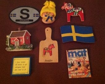 May include: A collection of Swedish themed magnets, including a red and white Dala horse, a yellow and blue Swedish flag, a red house, a wooden cutting board with a Dala horse, a yellow gnome, and a sticker with the letter "S" and the word "SVERIGE".