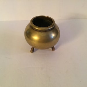 May include: A small, round, gold-coloured brass pot with three legs. The pot has a smooth, shiny surface.