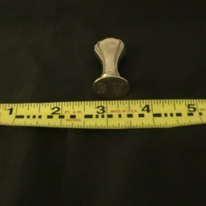 Op de afbeelding: Een zilverkleurige zegel met een decoratieve bovenkant en een platte basis. De basis is gegraveerd met de letters "K.Y.". De zegel wordt gemeten met een gele meetlint.