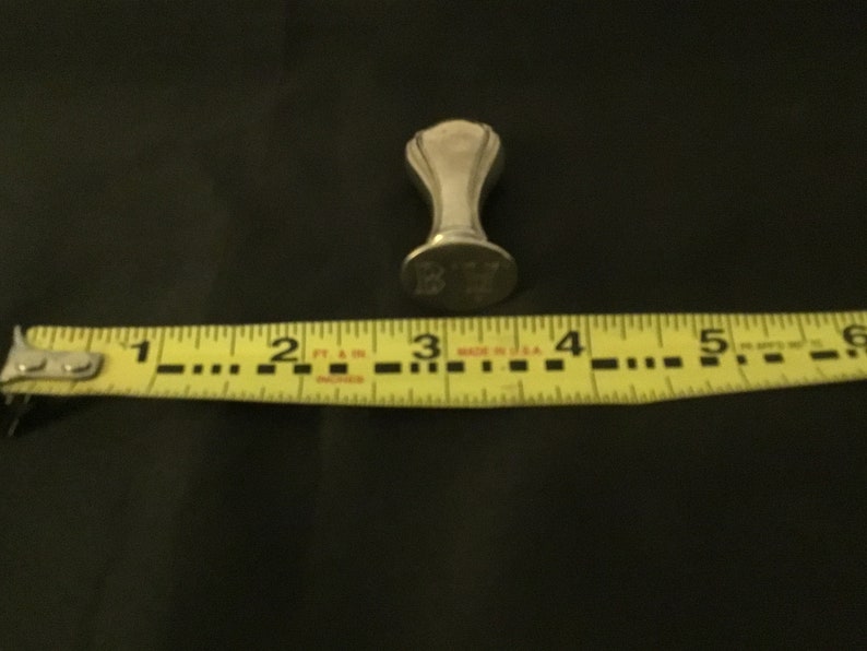 Op de afbeelding: Een zilverkleurige zegel met een decoratieve bovenkant en een platte basis. De basis is gegraveerd met de letters "K.Y.". De zegel wordt gemeten met een gele meetlint.