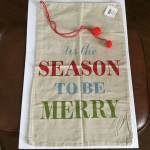 Può includere: Una borsa regalo in tela bianca con la scritta "'tis the season to be merry" in lettere rosse, blu e verdi. La borsa ha un cordoncino rosso con due pompon rossi.