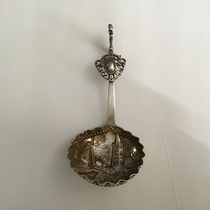May include: A silver tea strainer with a decorative handle and a bowl featuring an engraved image of a church and a ship.