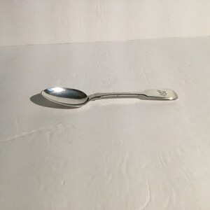 Puede incluir: Una cucharita de plata con un mango adornado. El mango tiene un diseño decorativo y está grabado con iniciales.