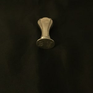 Op de afbeelding: Een zilveren zegel met een afgeronde bovenkant en een handvat. De zegel heeft de initialen "B.H." gegraveerd op de onderkant.